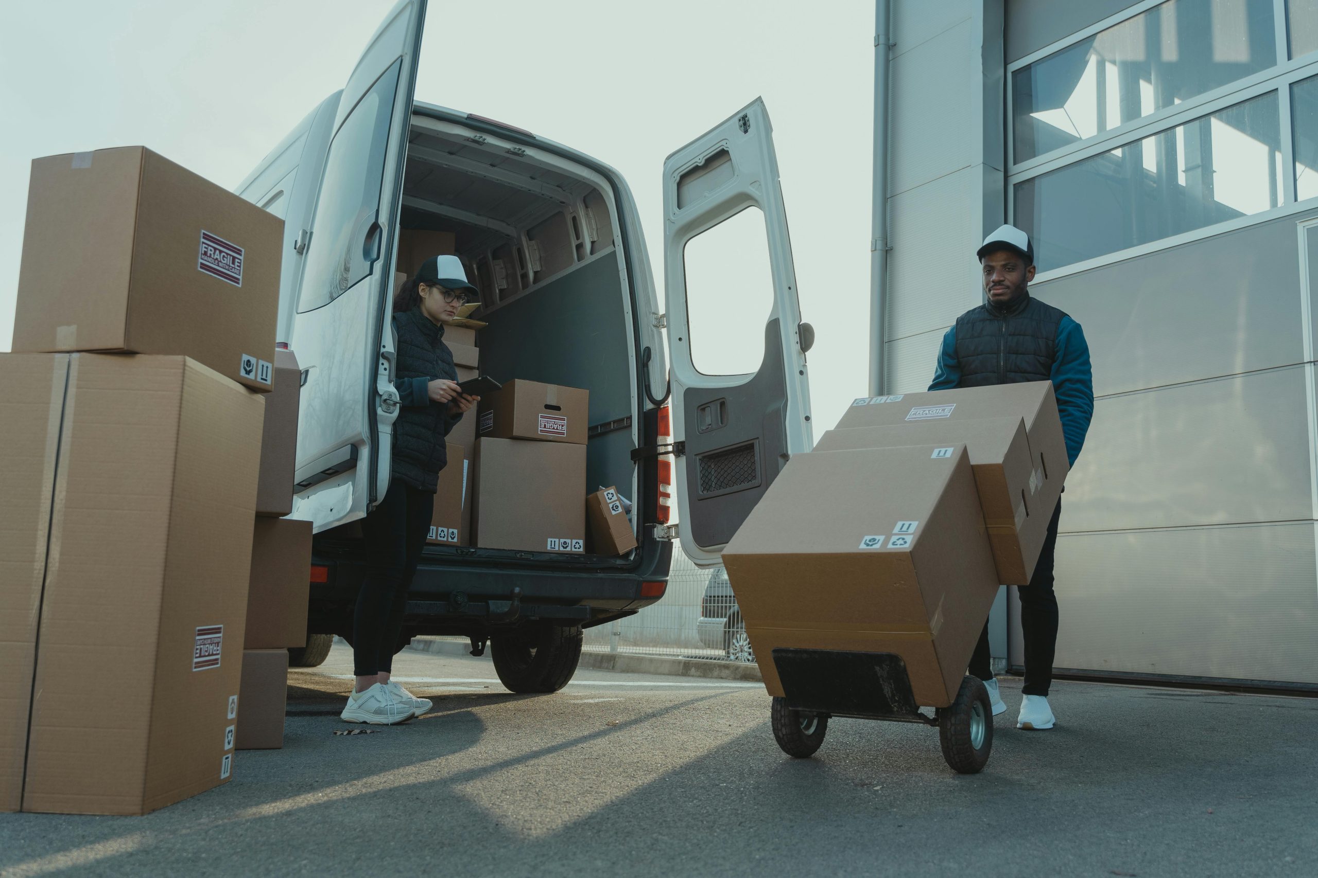 Loading van Two delivery workers unloading a white van outside a warehouse, handling multiple cardboard boxes labeled "Fragile." One worker is using a trolley to move boxes, while the other checks a clipboard, ensuring an organized and efficient delivery process.